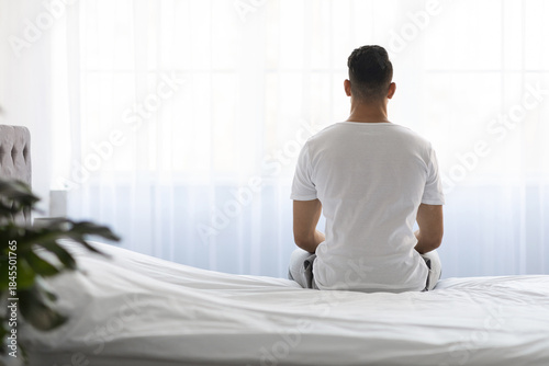 Фотография A young man in a white t-shirt sits on his bed with his back facing the viewer