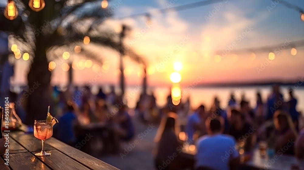 Naklejka premium Vibrant tropical cocktail with a garnish sits on a wooden bar counter while blurred people socialize at an outdoor beach club during a picturesque summer sunset