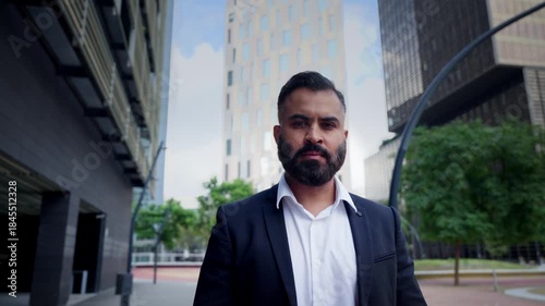 Confident Indian businessman in a suit stands in a modern business district, making direct eye contact at eye level. Urban skyline suggests leadership, corporate ambition and professional focus