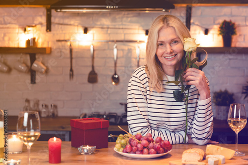 Adorable nice-looking middle-aged woman with rose sitting in the kitchen with romantic dinner on the table. Smiling lady celebrating special event birthday
