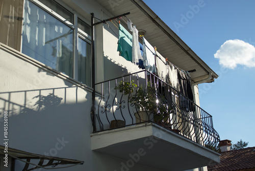 Residential balcony with iron railing and drying clothes under blue sky, strong shadows and copy space, for household, hygiene, textile care, everyday lifestyle and housing topics.