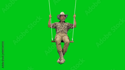 A middle aged man wearing camouflage with cowboy hat sitting on rope swing against on green screen