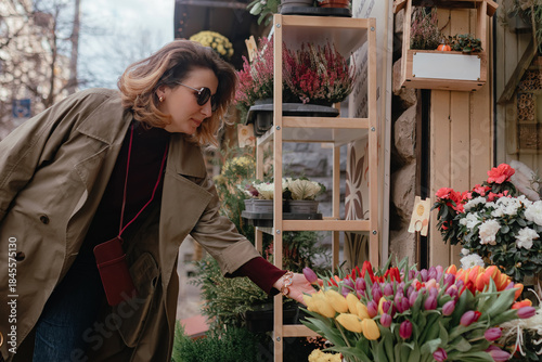 Fotografia Woman reaching toward tulip bouquet during floral stall visit, warm daylight, lively color palette, casual shopper movement