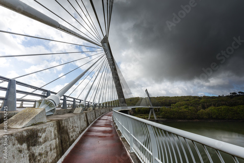 Menace de forte pluie sur un pont de forme courbe à haubans donc suspendu à des câbles, au-dessus d'une rivière. Pont de Térénez, France