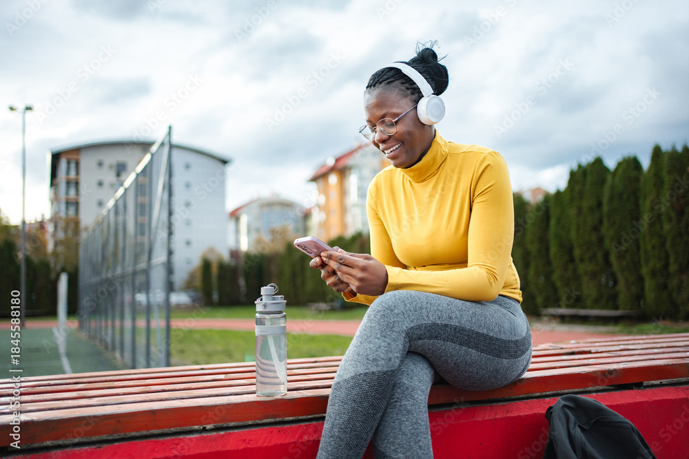 Obraz premium Young adult woman relaxing on bench using smartphone wearing headphones
