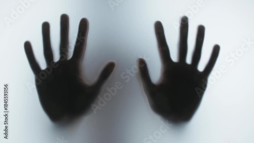 Dark blurry silhouette of two hands pressing against a frosted glass pane in a desperate, creepy, and scary scene