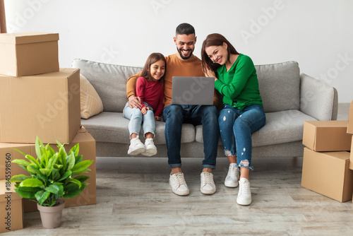 Foto A cheerful Middle Eastern family sits on a couch in their new flat, using a laptop to discuss furniture and decor while surrounded by moving boxes and a plant