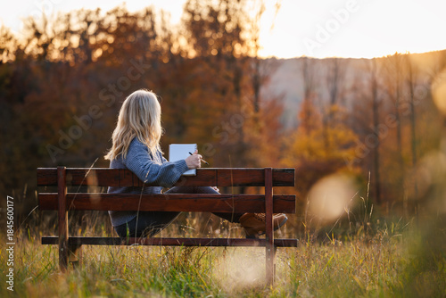 Relaxed woman sitting on park bench with notebook, quietly watching the sunset. Digital detox through journaling and diary writing