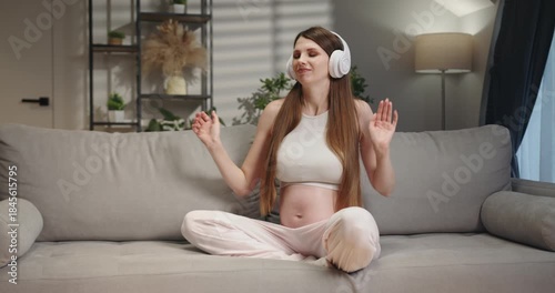 Pregnant woman wearing headphones sits on sofa, touching her belly with a smile, enjoying music and anticipating motherhood. Pregnant Woman Listening Music
