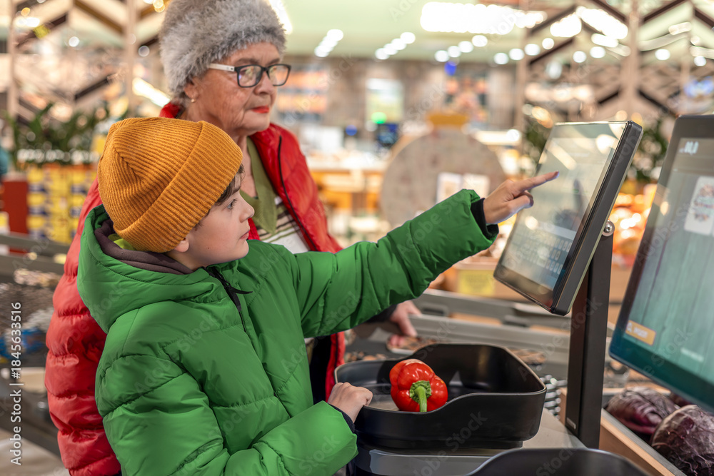 Obraz premium Young boy using self-checkout scale with grandmother supervision at grocery store produce section. modern retail experience, family bonding, digital literacy education and supervised independence