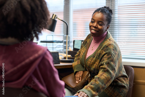Middle aged Black woman principal smiling and talking to student in office, sitting at desk with hand resting on student arm, engaging in supportive conversation during school meeting