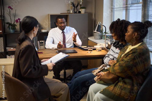 Bilde på lerret Middle aged Black man sitting at desk gesturing, while talking to two young Blac