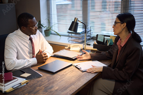 Foto Middle aged Black man sitting at desk listening to middle aged Caucasian woman d