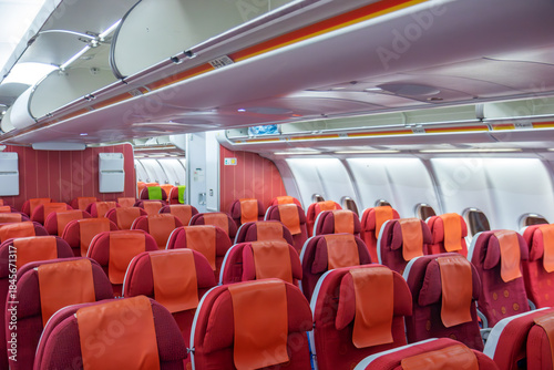 Interior of a passenger plane with empty red seats with orange headrests, overhead bins and bright cabin lighting.