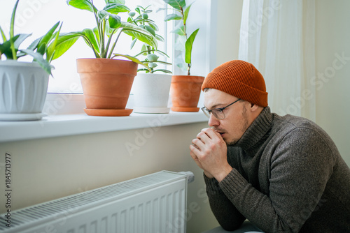 Man suffering from cold indoors due to high heating costs, keeping warm in a hat and sweater while trying to save money on expensive utilities, depicting energy crisis and home comfort issues