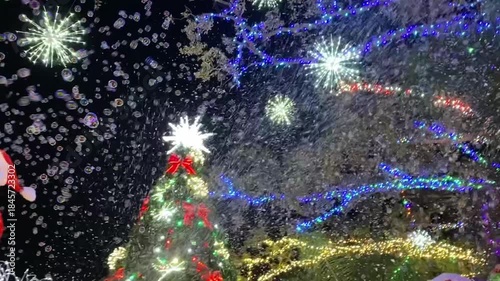 A close-up view shows an inflatable Santa with a candy cane and wreath beside a sparkling Christmas tree. Bubble-like snow and bright snowflake lights fill the dark night sky.