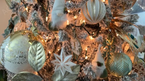 A decorated flocked Christmas tree features a silver poinsettia, green baubles, and white ornaments with warm lights. Elegant holiday decor creates a cozy seasonal mood.