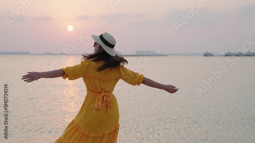 Smiling woman in a yellow dress twirling with open arms at the beach during golden sunset,

