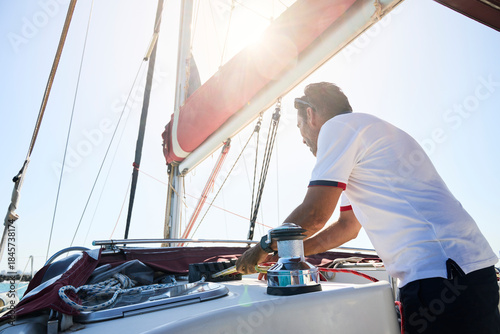Photos sailing instructor uses a powered winch to collect the ropes controlling the sails on a sailboat deck during sunset navigation