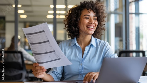 A smiling businesswoman with curly hair is reviewing documents at her desk with a laptop.