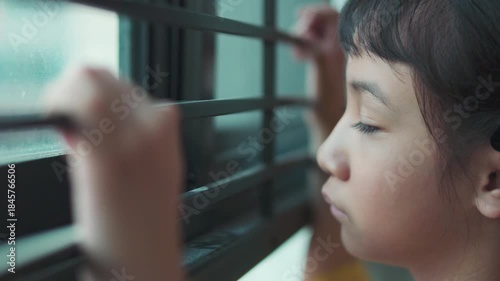 Close up side profile of sad Asian girl looking out window with selective focus. Lonely child face details with blurred hand holding bars in foreground, concept of waiting, depression, or boredom.