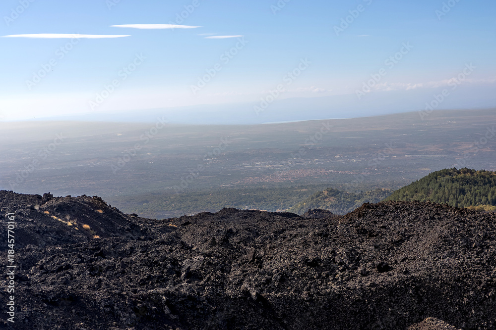 Obraz premium Volcanic landscape on the slopes of Mount Etna, Sicily, Italy. Dark solidified lava fields in the foreground with a panoramic view over the Sicilian countryside and distant coastline under a clear sky