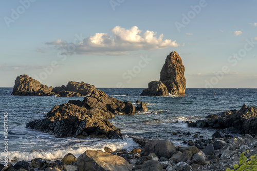 Volcanic lava rock formations rising from the Mediterranean Sea on the coast of Sicily, Italy. Rocky shoreline shaped by volcanic activity, with waves crashing against dark basalt rocks
