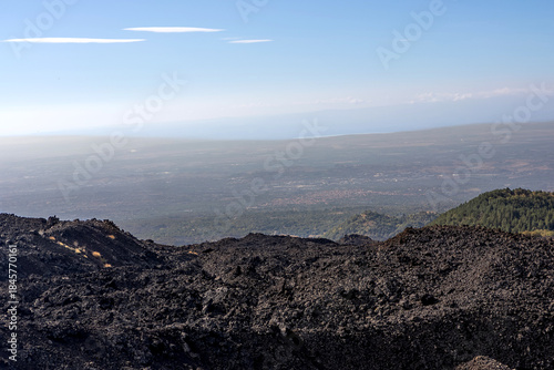 Volcanic landscape on the slopes of Mount Etna, Sicily, Italy. Dark solidified lava fields in the foreground with a panoramic view over the Sicilian countryside and distant coastline under a clear sky
