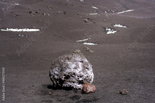 solitary volcanic rock resting on a dark lava field on Mount Etna, Sicily. The textured surface of hardened lava contrasts with scattered patches of snow in the background, creating a dramatic