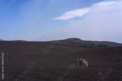 Wide volcanic landscape with dark lava fields and scattered rocks under a clear blue sky. Barren terrain of Mount Etna in Sicily creates a minimalistic, dramatic natural scene with strong sense