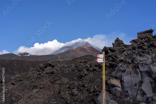 Volcanic hiking trail signpost standing among black lava rocks with Mount Etna visible in the background, partially covered by clouds. The image represents adventure travel, volcanic landscapes