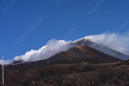 Summit of Mount Etna volcano rising above dark volcanic slopes, partially wrapped in white clouds under a clear blue sky. The image showcases the dramatic volcanic landscape of Sicily, Italy