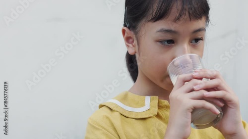Close up of adorable Asian girl drinking fresh milk from glass in natural light. Cute child holding milk glass enjoying healthy breakfast nutrition and calcium for growth and development.