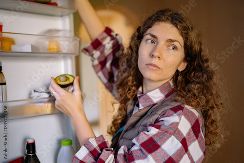 A woman with curly hair looks thoughtfully at an avocado as she stands in front of an open refrigerator in a kitchen. She seems to decide on a meal