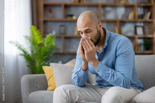 Tableau sur toile Young man using a tissue to blow his nose while sitting on a couch at home, expe