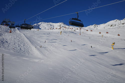 A scenic winter landscape at a modern alpine ski resort featuring chairlifts, skiers, and snow-covered mountain slopes under a clear blue sky.