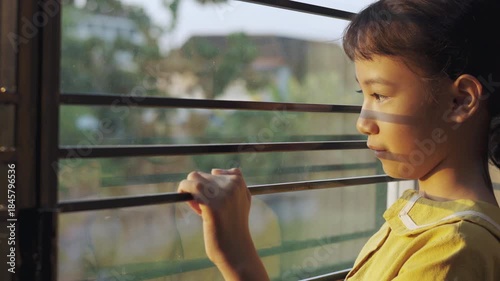 Sad Asian girl looking out window through iron bars during sunset. Lonely child waiting with hope, sun shadows on face and reflection on glass. Concept of depression, solitude, or home isolation.