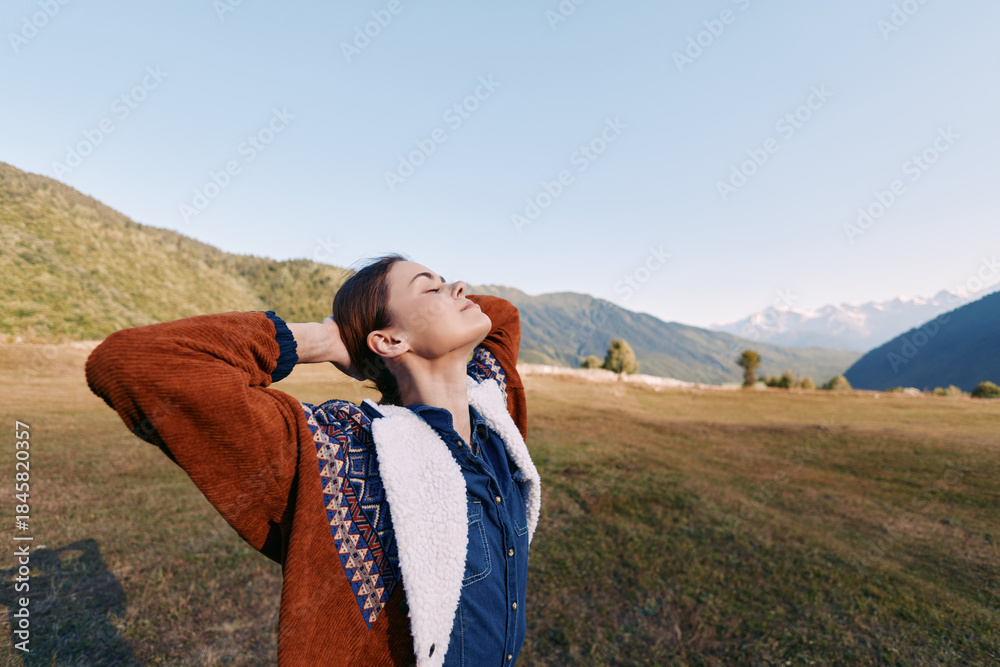 Fototapeta premium Woman in nature among mountains and a wide field, enjoying relaxation and freedom outdoors during a travel break, eyes closed and arms raised in a peaceful autumn landscape.