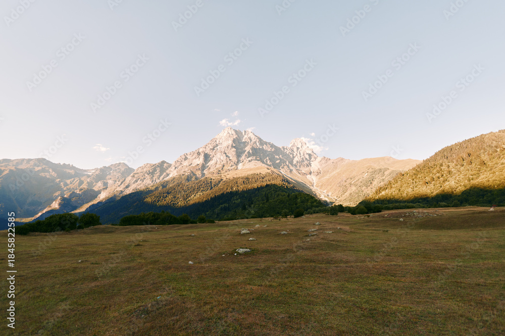 Fototapeta premium Mountain meadow landscape with a towering peak under clear sky, alpine grassland bathed in warm sunlight, panoramic vista of rolling hills and tranquil natural scenery at dawn.