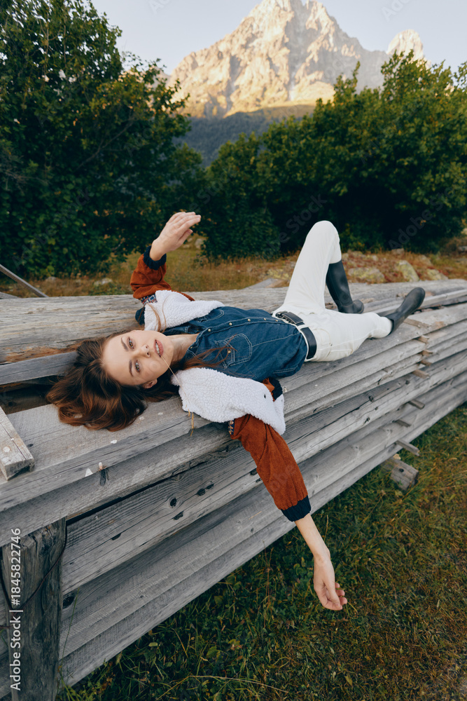 Fototapeta premium Woman model lying on wooden bench outdoors near mountain, fashion outfit and relaxed pose. Nature scene with trees, grassy meadow and casual editorial portrait in alpine setting.