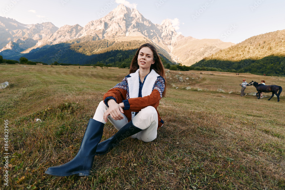 Fototapeta premium Woman sits in mountain meadow wearing sweater and boots, enjoying countryside nature with horses grazing in background, autumn light and relaxed outdoor lifestyle portrait.