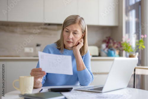 Mature woman analyzing financial documents and bills, calculating expenses, and managing stressed personal accounting while sitting at a kitchen table with a laptop