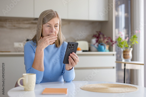 Canvas Print Senior woman in kitchen covers her mouth in shock and worry while reading upsett