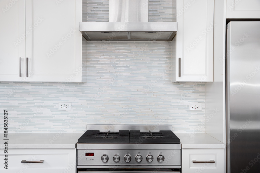 Fototapeta premium A modern white kitchen features a stainless steel gas stove against a light blue and white mosaic tile backsplash. 