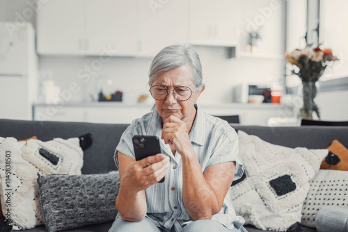 Fotografie Elderly woman sits on a sofa at home looking at her phone with disappointment