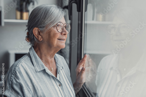 Elderly woman stands by the window smiling while looking outside. She enjoys peaceful leisure moments and dreams positively about her future at home.