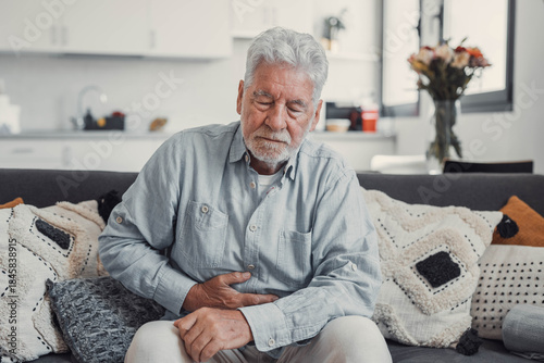 Elderly man sits on a sofa holding his stomach in pain. He struggles with abdominal discomfort and fatigue while trying to relax at home.