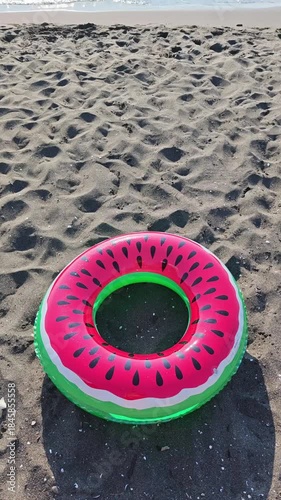 Vibrant, inflatable swim ring with a watermelon design resting on the sandy shore of a beach, with calm ocean waves gently rolling in the distance