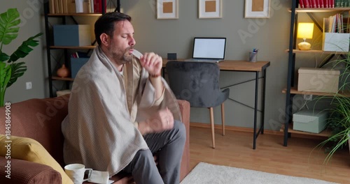 Side view of sick man coughing on couch covered with cozy blanket