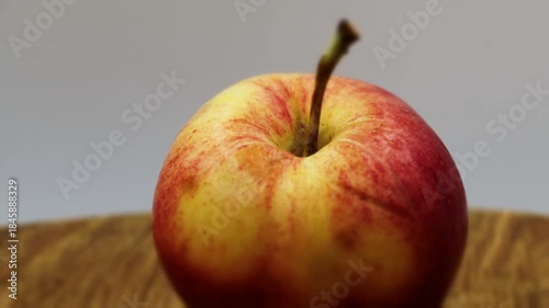A close-up view of a single, vibrant red and yellow apple, covered in sparkling water droplets, slowly rotating on a rustic wooden cutting board. 